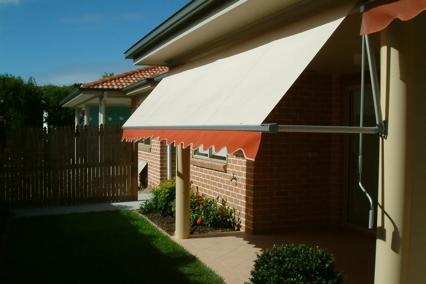 Cream pivot arm awning with an orange valance angled out over a window beside a lawn