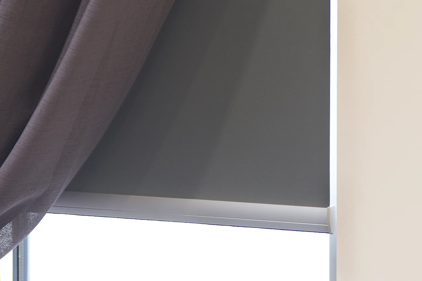 Charcoal blockout roller blind lowered behind a sheer curtain, with light only at the bottom edge