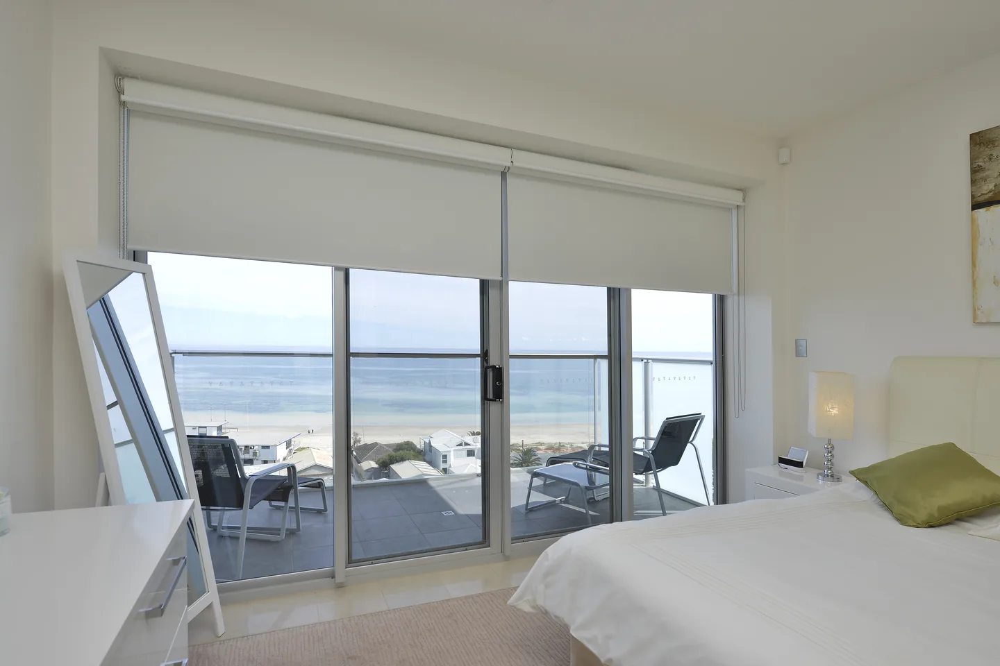 Bedroom opening to a balcony and beach view, with roller blinds rolled up at the head of the sliding doors