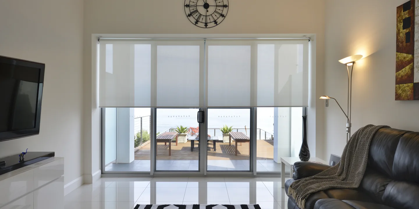 Living room with light sunscreen roller blinds lowered part way across wide sliding doors to a coastal deck