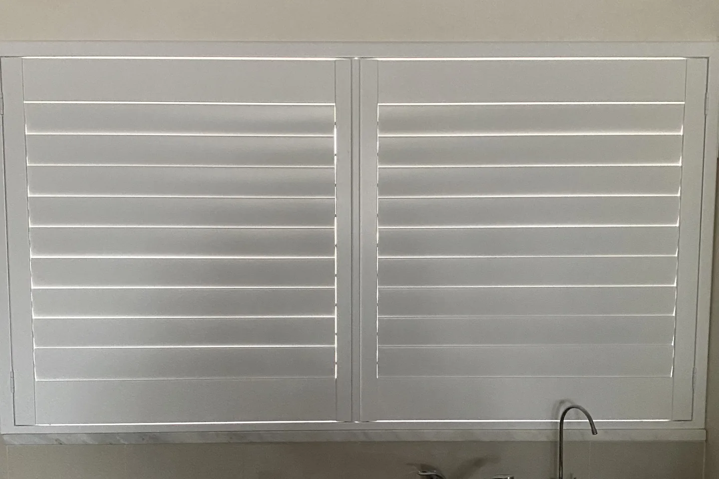 White plantation shutter panels over a kitchen window with the louvres tilted closed, light showing only along the blade edges
